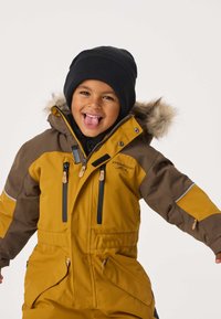 Geel en bruin geïsoleerd snowsuit met een capuchon met bontrand, meerdere ritsen en een relaxed fit. Zwarte gebreide beanie op het hoofd.