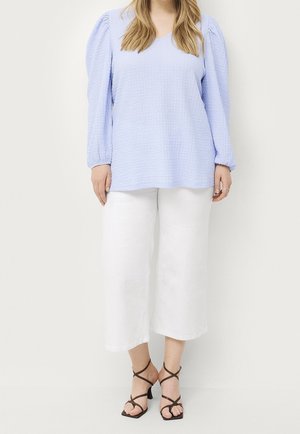 Femme portant un haut bleu clair texturé à manches longues, un pantalon blanc large et court, et des sandales à talons noires avec des lanières, debout devant un fond blanc.
