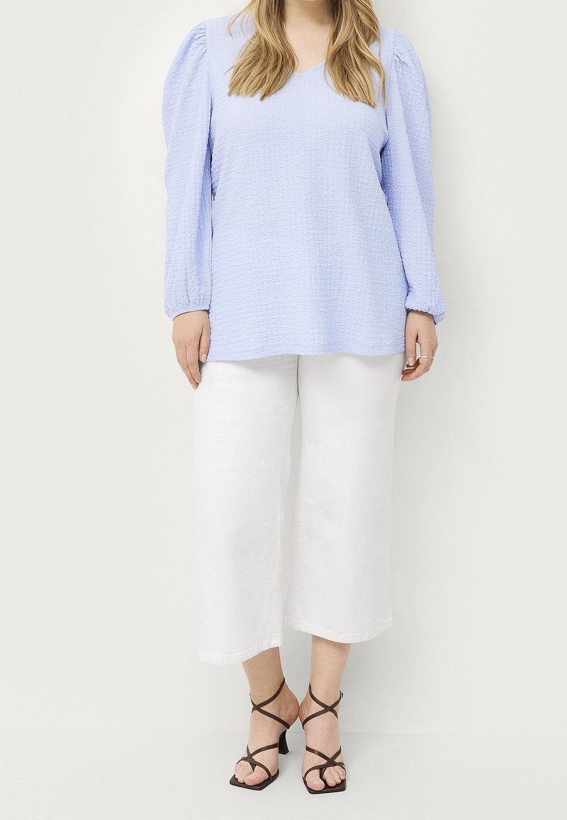 Femme portant un haut bleu clair texturé à manches longues, un pantalon blanc large et court, et des sandales à talons noires avec des lanières, debout devant un fond blanc.