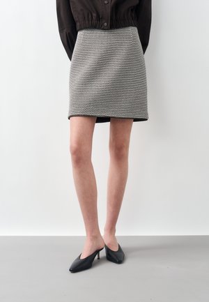 Model purtând o mini-fustă în carouri alb-negru, o jachetă scurtă neagră și mule negre cu toc și vârf ascuțit, stând pe un fundal alb.