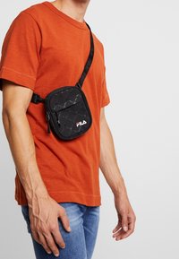 Czarna torba na ramię Fila z fakturowanym wzorem, zamykana na zamek, z regulowanym paskiem. Nosi się ją na pomarańczowej koszuli na osobie stojącej.