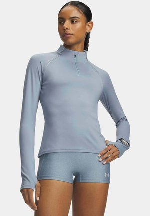 Mujer con cabello trenzado que lleva una camiseta deportiva de manga larga azul claro y pantalones cortos azules, posando con una mano en la cadera y reloj inteligente visible.