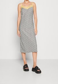 Robe midi à fleurs noir et blanc avec bordure en dentelle, bretelles jaunes et coupe ajustée. Stylisée avec des sandales noires épaisses.