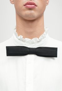 Černý motýlek z hladké látky, umístěný na bílé košili s volánovým límcem. Motýlek má klasický, plochý tvar.