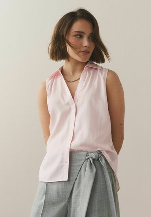 Kobieta o krótkich brązowych włosach, ubrana w bezrękawową, jasnoróżową koszulę na guziki, wpuszczoną w wysokowalne szare spodnie z przewiązanym paskiem w talii.