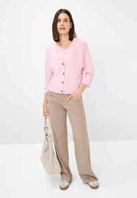 Light pink cardigan with wooden buttons, V-neck, three-quarter sleeves, paired with beige wide-leg pants and a woven beige bag.