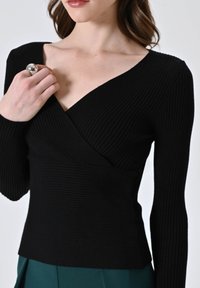 Top lungo a maniche nere a coste con scollatura a V e design avvolgente, caratterizzato da un tessuto testurizzato e una vita aderente. Modella che tiene un anello.