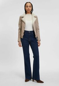 Beige faux leren jas met een schapenvacht kraag, gecombineerd met hooggesneden, flare blauwe jeans. Het model draagt een witte getailleerde top en bruine hakken.