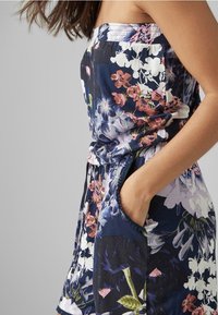 Jumpsuit floral con base azul marino, presentando patrones en rosa, blanco y verde. Incluye un escote elasticado y bolsillos laterales.
