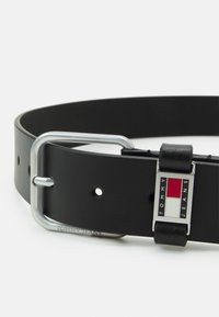 Zwarte leren riem met een zilveren metalen gesp. De gesp heeft een rechthoekig logo met rode en witte strepen en gegraveerde tekst.