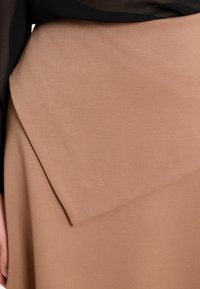 Brauner, strukturierter Rock mit einem gefalteten Klappendetail. Der Stoff wirkt weich mit einer glatten Oberfläche und ergänzt ein durchscheinendes schwarzes Oberteil.