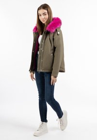 Parka verde oliva con cappuccio in pelliccia sintetica rosa, dotata di zip frontale, bottoni a pressione, tasche laterali e vestibilità rilassata. Abbinata a jeans blu e sneakers bianche.
