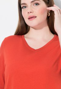 Maglietta rossa con scollo a V e maniche corte, realizzata in tessuto morbido. Presenta una vestibilità ampia e una trama liscia. I dettagli includono cuciture sottili.