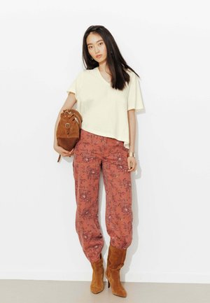 Femme portant un haut crème à manches courtes, un pantalon floral couleur rouille, des bottes en daim beige, tenant un sac à main assorti en daim beige sur fond blanc.