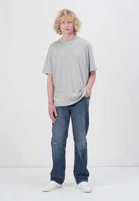 Graues Baumwoll-T-Shirt mit relaxed Fit und kurzen Ärmeln, kombiniert mit blauen Jeans im geraden Schnitt und leichter Zerrung. Weiße Sneaker.
