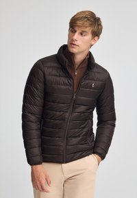 Brun vadderad jacka med hög krage, med horisontellt kviltat mönster och dragkedjestängning. Accentuerad med en liten logotyp på bröstet.