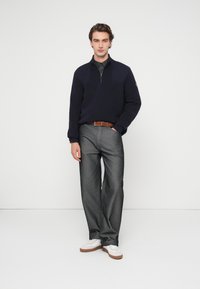 Pull-over en maille bleu marine avec une fermeture à glissière sur le devant, accompagné d'un pantalon gris à jambes larges et de baskets blanches avec des semelles marron. Ceinture en cuir marron incluse.