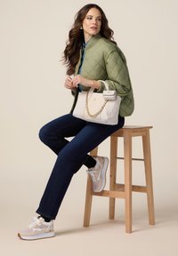 Chaqueta acolchada verde oliva, camisa de mezclilla azul, pantalones oscuros y zapatillas beiges, sosteniendo un bolso blanco con cadena dorada, sentado en un taburete de madera.