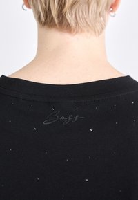 Melnas t-kreklas ar noapaļotu kakla izgriezumu, kura priekšpusē ir tekstūrēta logotipa izšuvums tumšā diegā un nelielas spīdīgas akcenti visā audumā.