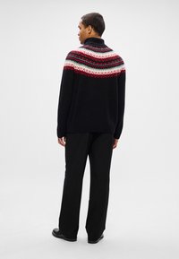Homme portant un pull noir à col roulé avec un yoke à motifs rouge, blanc et gris, un pantalon noir et des chaussures noires, se tenant devant un fond uni.