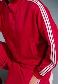 Personne portant une veste rouge zippée et un pantalon assorti avec trois bandes blanches sur les manches et les jambes, main dans la poche, assise ou accroupie.