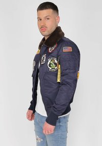 Marineblaues Bomberjackett mit einem Fellkragen, mehreren Aufnähern, Reißverschluss vorne, gerippten Bündchen und einem Schlüsselanhänger. Getragen über einem weißen Shirt und Jeans.