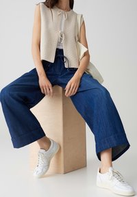 Femme assise sur un bloc de bois portant un gilet en tricot beige sans manches, un jean large bleu, des baskets blanches et un sac cabas beige à l'épaule.
