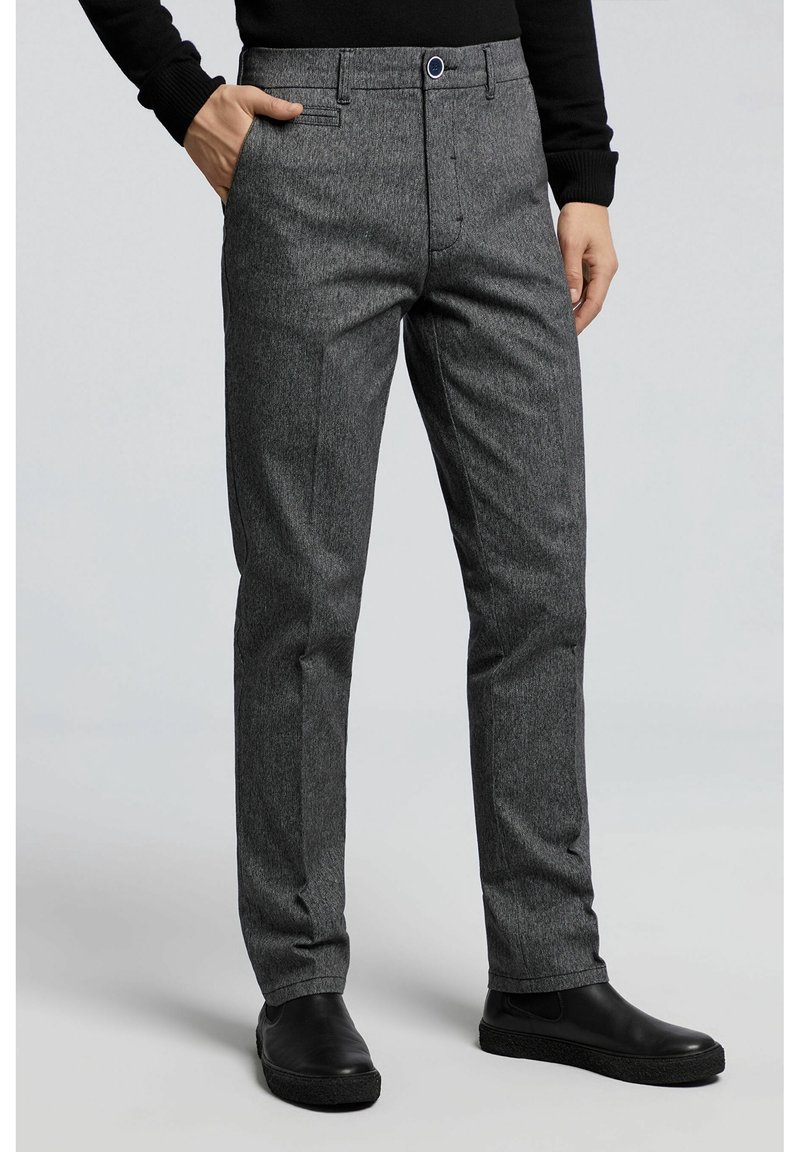 Homme portant un pantalon gris cintré avec poches latérales et des chaussures noires sans lacets, debout devant un fond gris uni.
