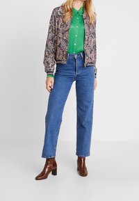 Mønsteret bomberjakke med paisley-design, grøn skjorte med knapper, højtaljede jeans og brune hælede støvler i krokodillemønster.