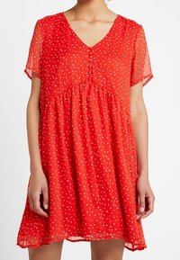 Robe courte rouge à manches courtes avec un col en V, dotée d'une fermeture boutonnée à l'avant, d'une coupe ample, et de motifs à pois blancs sur un tissu transparent.