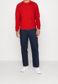 Roter Strickpullover mit rundem Ausschnitt, kombiniert mit dunkelblauen Jeans mit geradem Bein und weißen Sneakers. Schlichtes Design und einfarbige Farben.