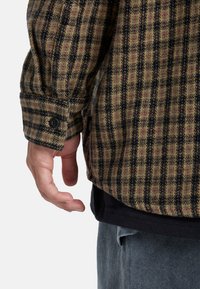 Camisa de franela a cuadros en oliva, negro y rojo. Presenta mangas largas con puños abotonados y un dobladillo curvado. El material parece tener textura.
