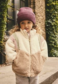 Enfant portant une veste en fourrure crème avec des segments en blocs de couleur rose clair, vert et beige. Accompagnée d'un bonnet en côtes violet.