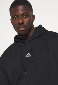 Zwarte hoodie gemaakt van zachte stof, met een wit Adidas-logo op de borst, trekkoorddetails en een relaxte pasvorm.