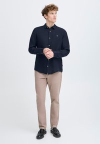 Camicia navy con maniche lunghe e polsini a bottoni, abbinata a chinos beige e scarpe nere. La camicia presenta un piccolo logo sul petto.