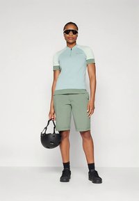 Svetlo modra športna majica s kratkimi rokavi in zadrgo, kombinirana z zelenimi kolesarskimi kratkimi hlačami. Kolo čelada v eni roki, vidni črni čevlji.