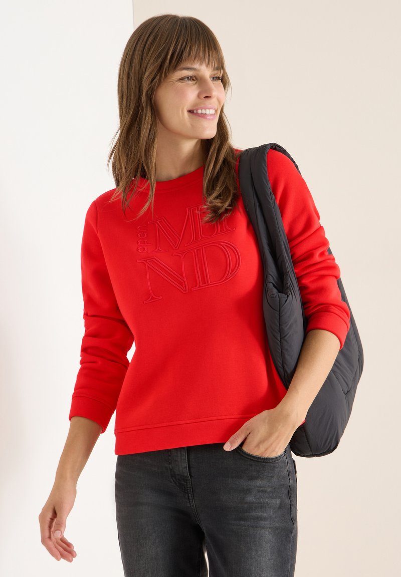 Sudadera roja con el texto en relieve "MENTES ABIERTAS", cuello redondo y mangas largas, combinada con jeans negros y un bolso acolchado oscuro.