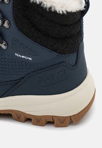 Detailní záběr na patu námořnicky modré turistické boty s textilní látkou Texapore, smetanovým mezipodešvem, hnědou robustní podešví a huňatým černobílým límcem.