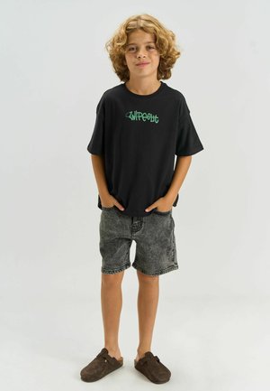 Poiss lokkis juustega seisab käed taskutes, seljas must "Wipeout" t-särk, pestud mustad teksapüksid ja pruunid sussitaolised kingad valgel taustal.