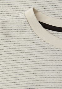 Zbliżenie kremowej koszulki z dekoltem w łódkę, cienkimi czarnymi poziomymi paskami i czarną wewnętrzną metką kołnierza.