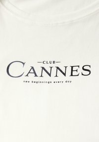 Bílý tričko s velkým černým potiskem, na kterém je napsáno "CLUB CANNES" a pod ním menším písmem fráze "nové začátky každý den".