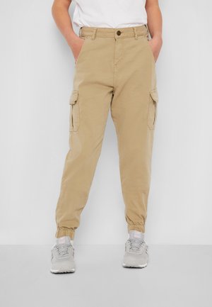 Person iført beige cargobukser med elastiske manchetter, hvide sneakers og en hvid skjorte, stående med hænderne i lommerne.