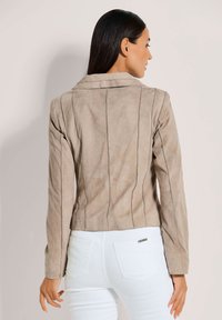 Beige Wildlederjacke mit vertikalen Nahtdetails und einem Reverskragen, von hinten angesehen, kombiniert mit weißen Hosen.