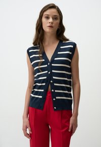 Marineblauwe mouwloze cardigan met witte horizontale strepen, V-hals en zilveren knopen. Gecombineerd met rode hoge broeken.