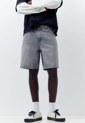 Osoba oblečená v sivých riflových kraťasoch, bielych ponožkách a bielych teniskách s čiernymi podrážkami a šnúrkami, stojaca so založenými rukami na bielom pozadí.