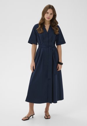 Femeie cu păr lung și castaniu, purtând o rochie midi albastru închis, cu curea, și sandale cu toc negre, stând pe un fundal simplu.