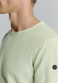 Lichtgroen shirt met korte mouwen van textuurstof, met een ronde halslijn en een hexagonaal logo-accent op de linkerschouder.