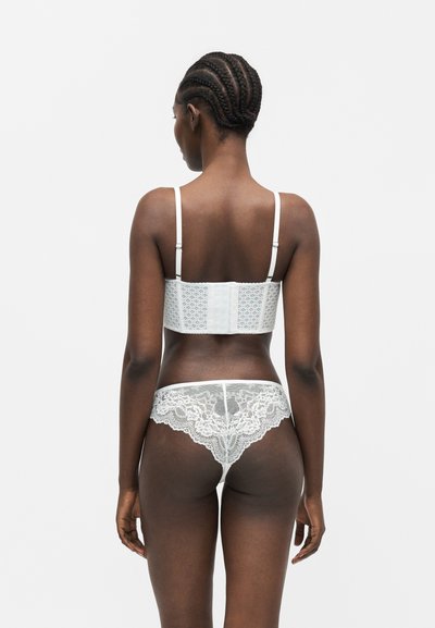 Γυναίκα με πλεξούδες στα μαλλιά, φοράει λευκό δαντελένιο μακρύ σουτιέν και ταιριαστό δαντελωτό σλιπ, εμφανιζόμενη από πίσω σε λευκό μονόχρωμο φόντο.