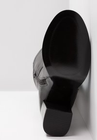 Botte en cuir noir inclinée vers le haut montrant une semelle texturée détaillée et un talon empilé sur fond blanc.