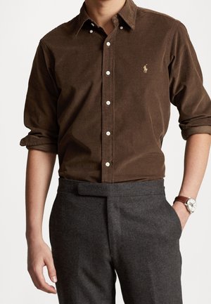 Homme portant une chemise marron à boutons avec les manches retroussées, un pantalon gris, et une montre-bracelet avec un bracelet en cuir marron.